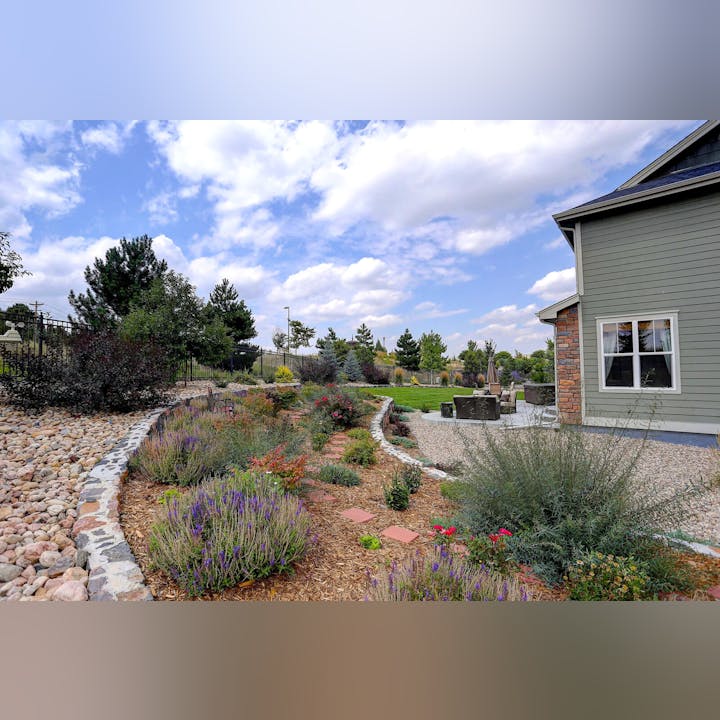 A landscaped garden with colorful plants and a stone path next to a house. There's outdoor seating on a patio under a partly cloudy sky.