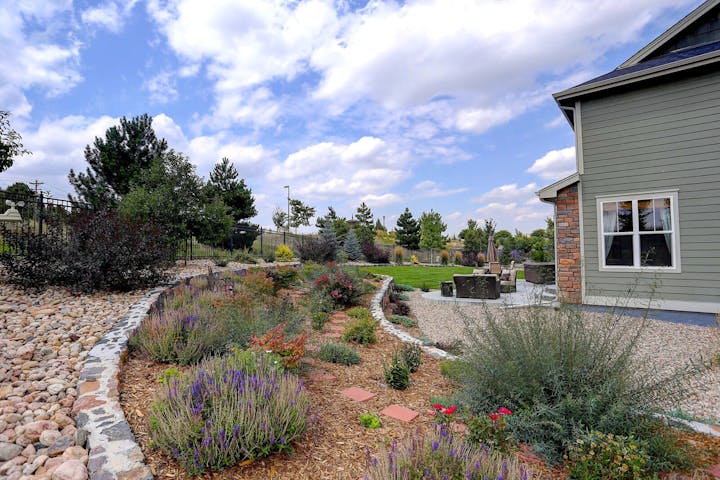 A landscaped garden with colorful plants and a stone path next to a house. There's outdoor seating on a patio under a partly cloudy sky.