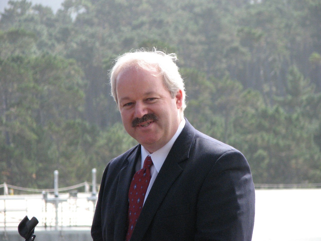 A man in a suit with a red tie smiling outdoors, trees and a fence in the background.