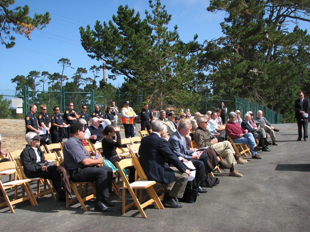 Outdoor event with people seated, a group standing, and a man addressing the audience under a clear sky.