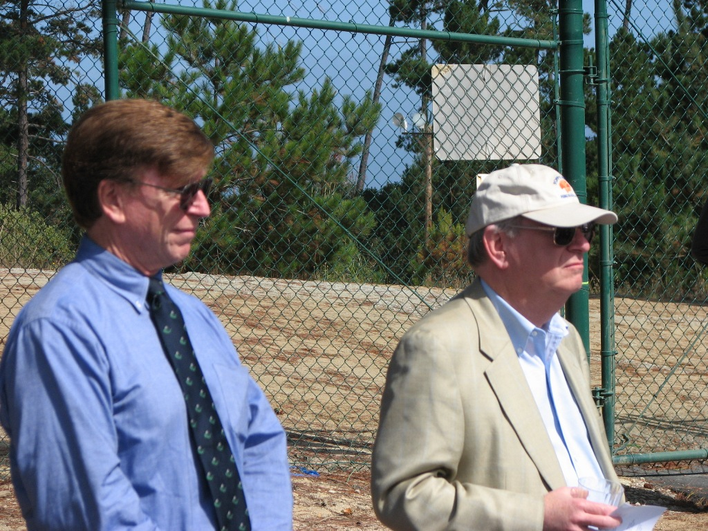 Two men by a fence with trees behind them, one wearing a cap.