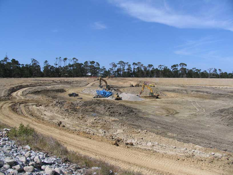 Construction site with excavators, clear sky, dirt tracks, and sparse trees.