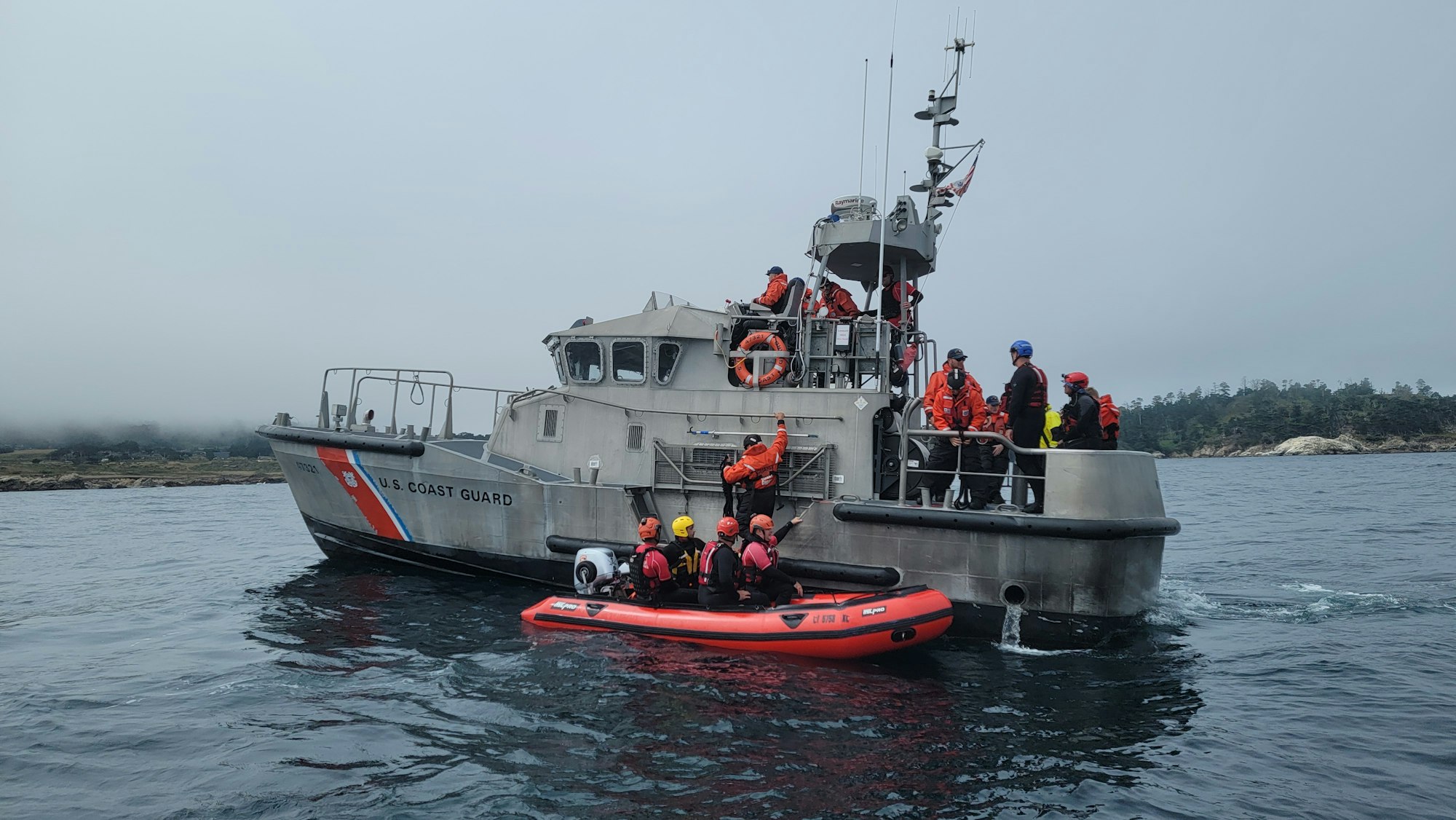 Coast Guard personnel in orange gear on a boat with a smaller rescue boat nearby, on a foggy day.