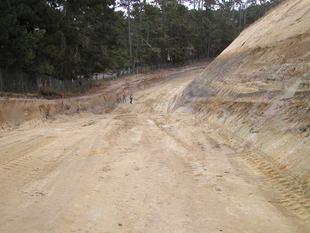 A dirt track with three people walking, surrounded by excavated earth and trees in the background.