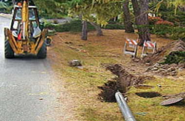 A digger beside a trench near a road with a barrier and downed guardrail.