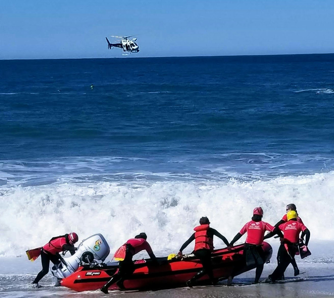 Five people launching a red rescue boat into the ocean, with a helicopter hovering above.