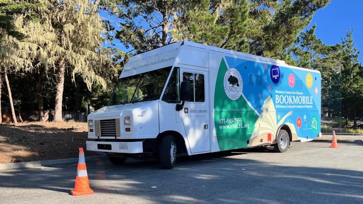 A colorful bookmobile from the Monterey County Free Libraries parked on a sunny day with trees in the background.