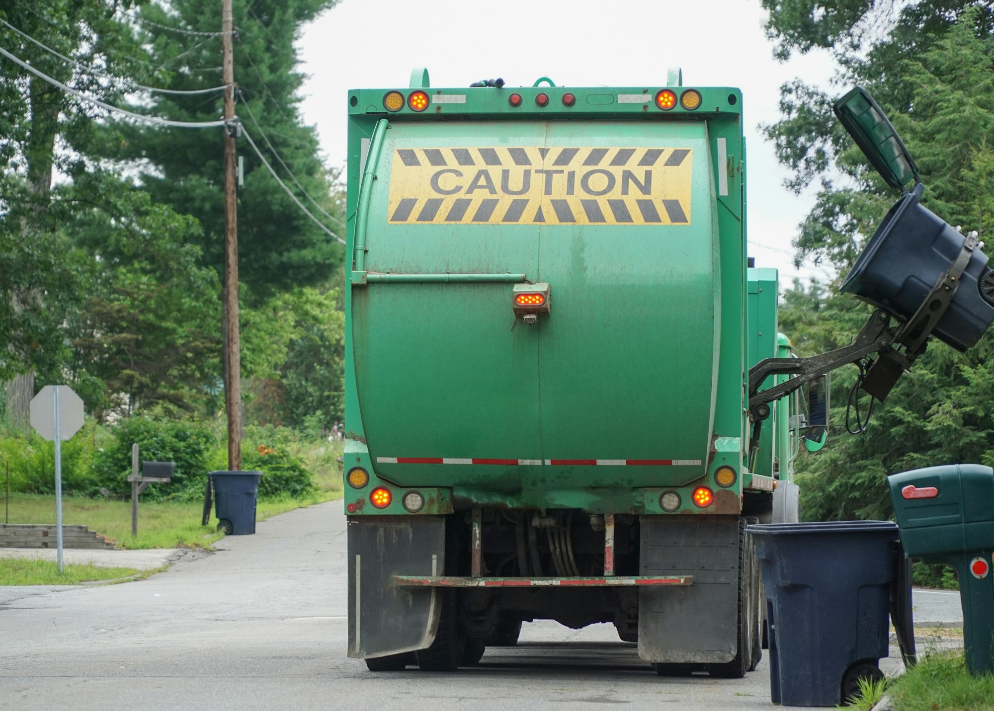 A green garbage truck with a "CAUTION" sign is parked on a street, while a trash can is being emptied into it.