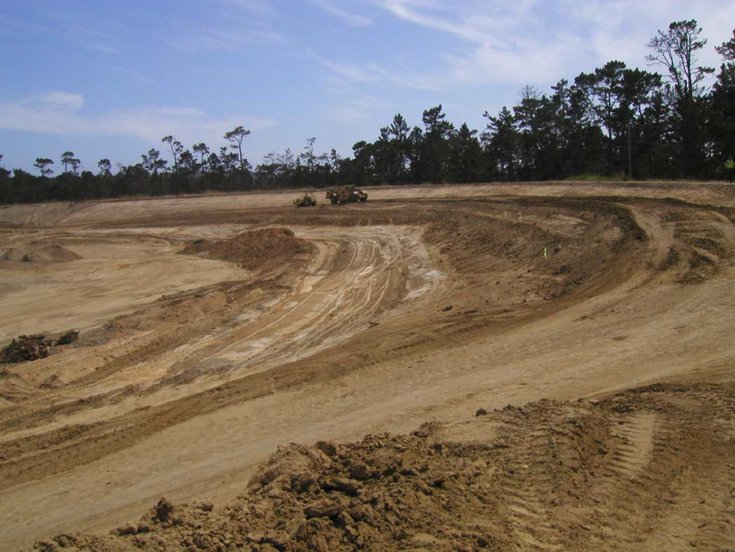 A construction site with exposed soil and machinery, surrounded by trees under a clear sky.