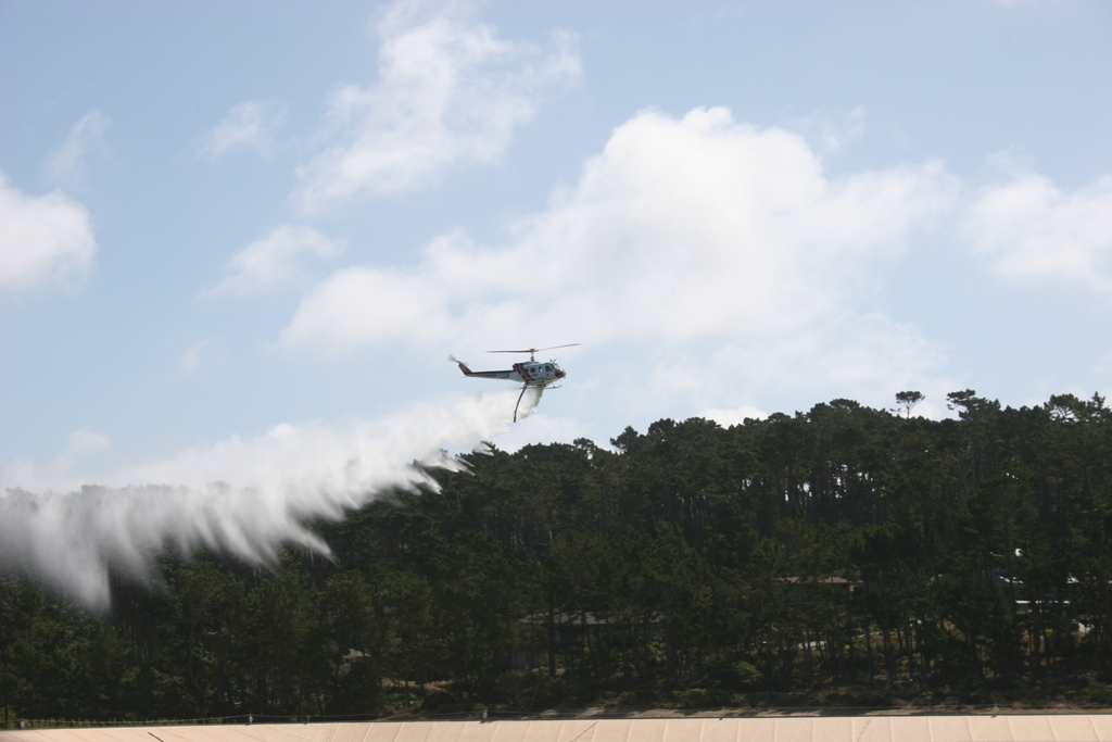 Helicopter spraying water above trees against a blue sky.