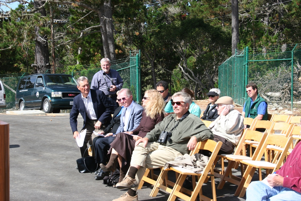 Group of people sitting on folding chairs outdoors, some chatting, others waiting, with trees and a van in the background.