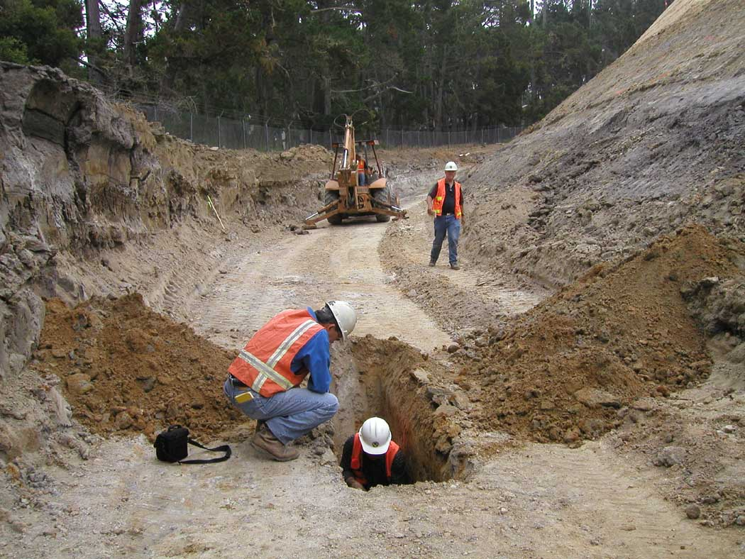 Construction site with workers, an excavator, and a deep trench in a dirt area.