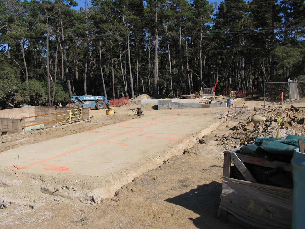 A construction site with a concrete foundation, heavy machinery, and a wooded background.