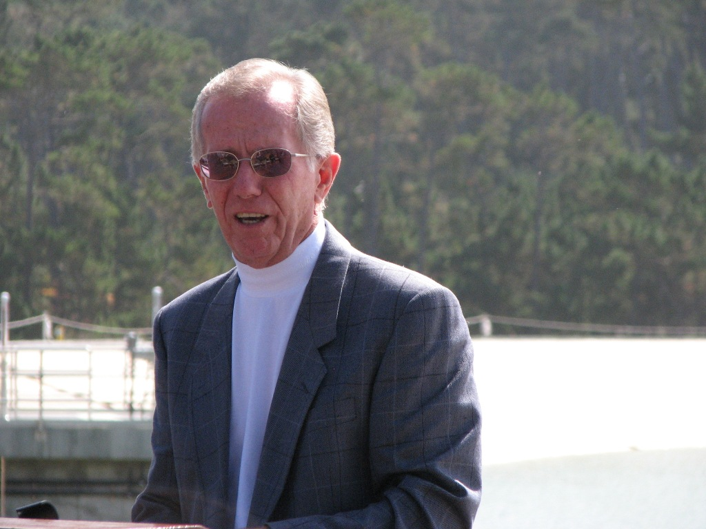 An older man wearing sunglasses and a suit, standing outdoors with a blurry green background.