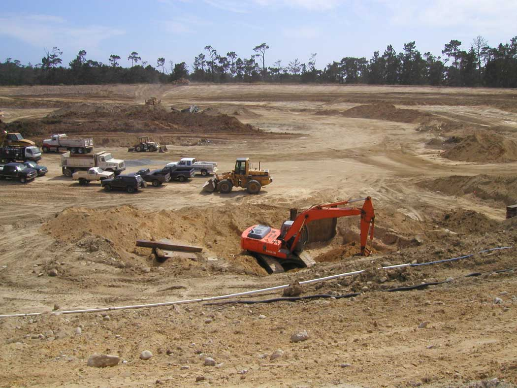 Construction site with excavator, trucks, and scattered equipment on dirt terrain.