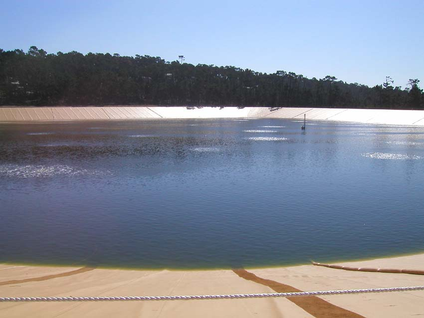 A water reservoir with a covered slope, surrounded by trees under a clear sky.