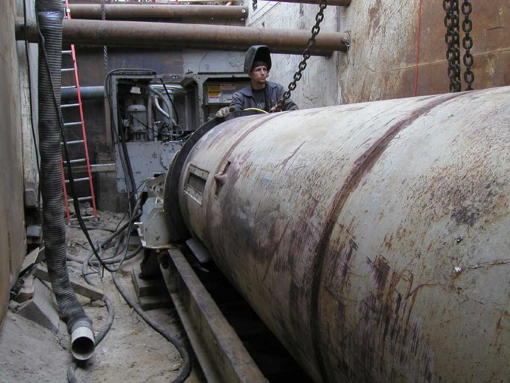 A worker beside a large cylindrical structure, possibly machinery, with chains and industrial equipment around.