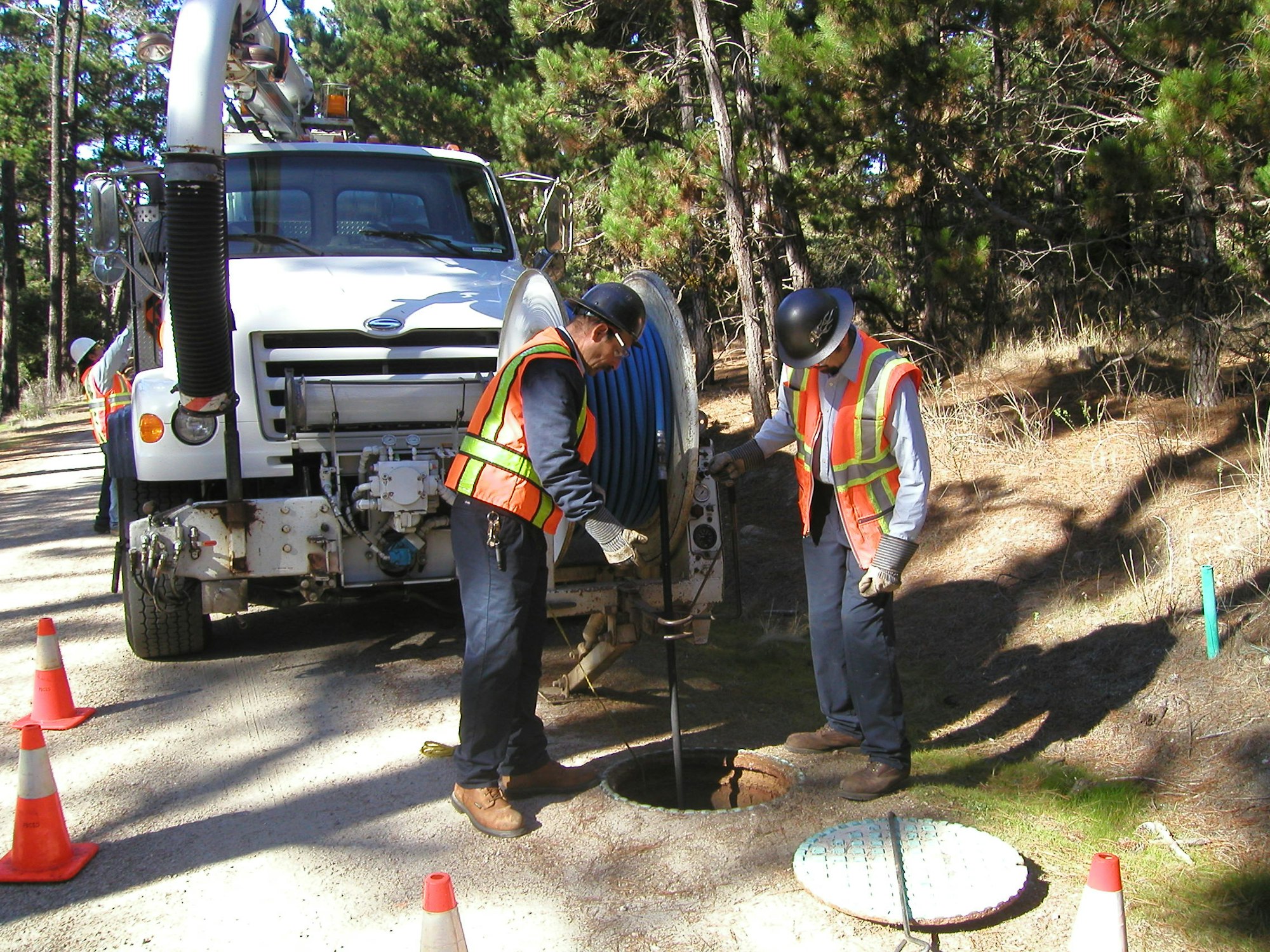 Two workers in safety gear using a truck-mounted drill near a manhole, with traffic cones for safety.
