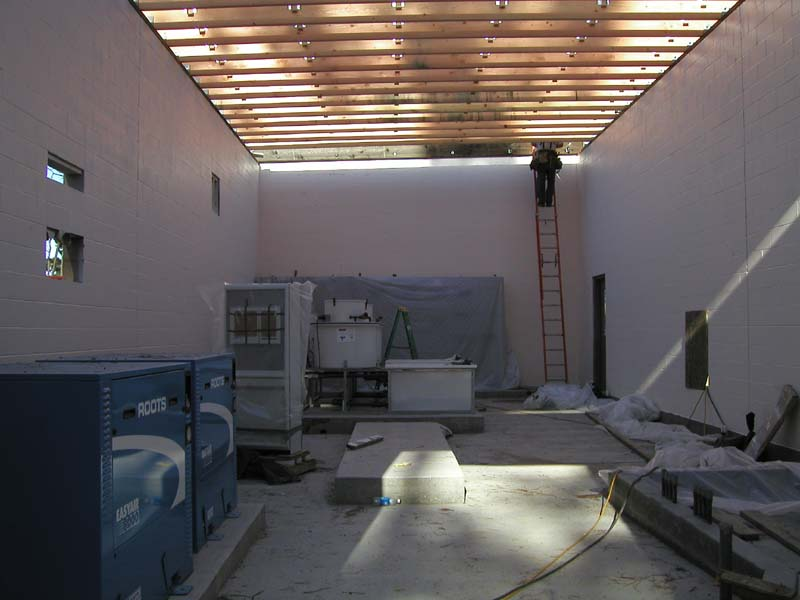 Industrial equipment in a room under construction with a person on a ladder and sunlight through the slatted roof.