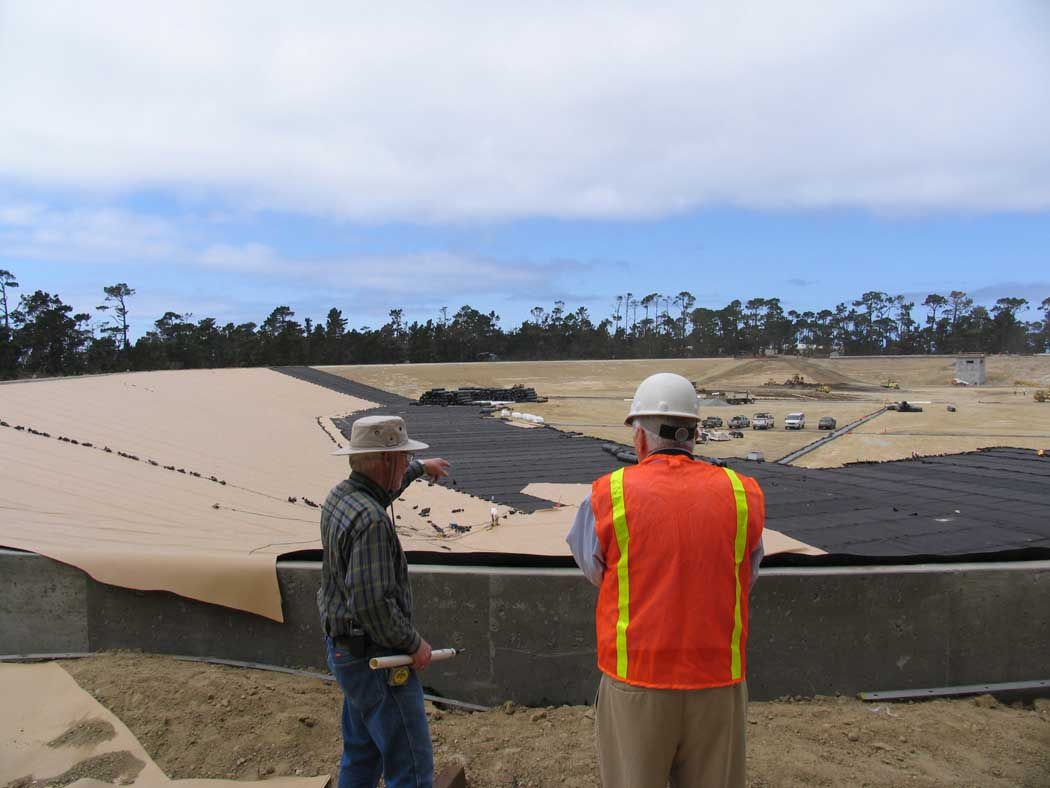 Two men observing a construction site with covered sand dunes and equipment under a cloudy sky.