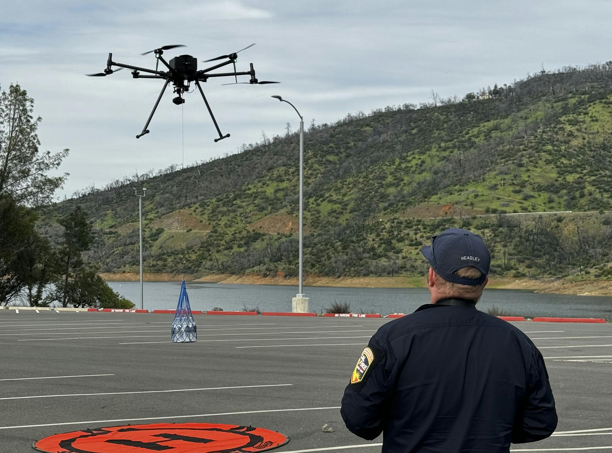 A person in uniform operates a drone outdoors near a marked landing pad, with hills and overcast skies in the background.