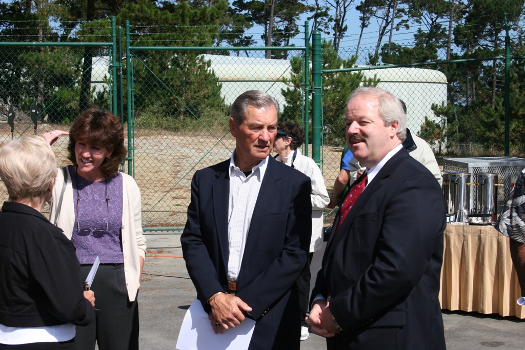 People gathered outdoors, two men in suits in foreground, one chatting, casual event vibe, trees and fencing in background.