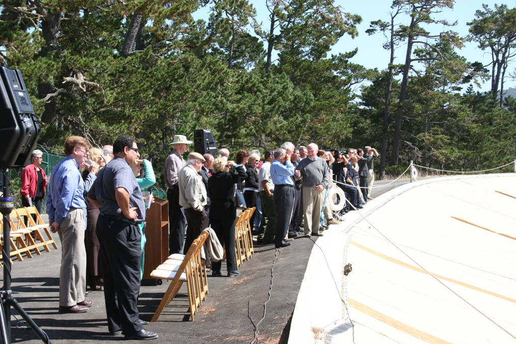 Group of people at an outdoor event with speakers, chairs, and audio equipment, surrounded by trees.