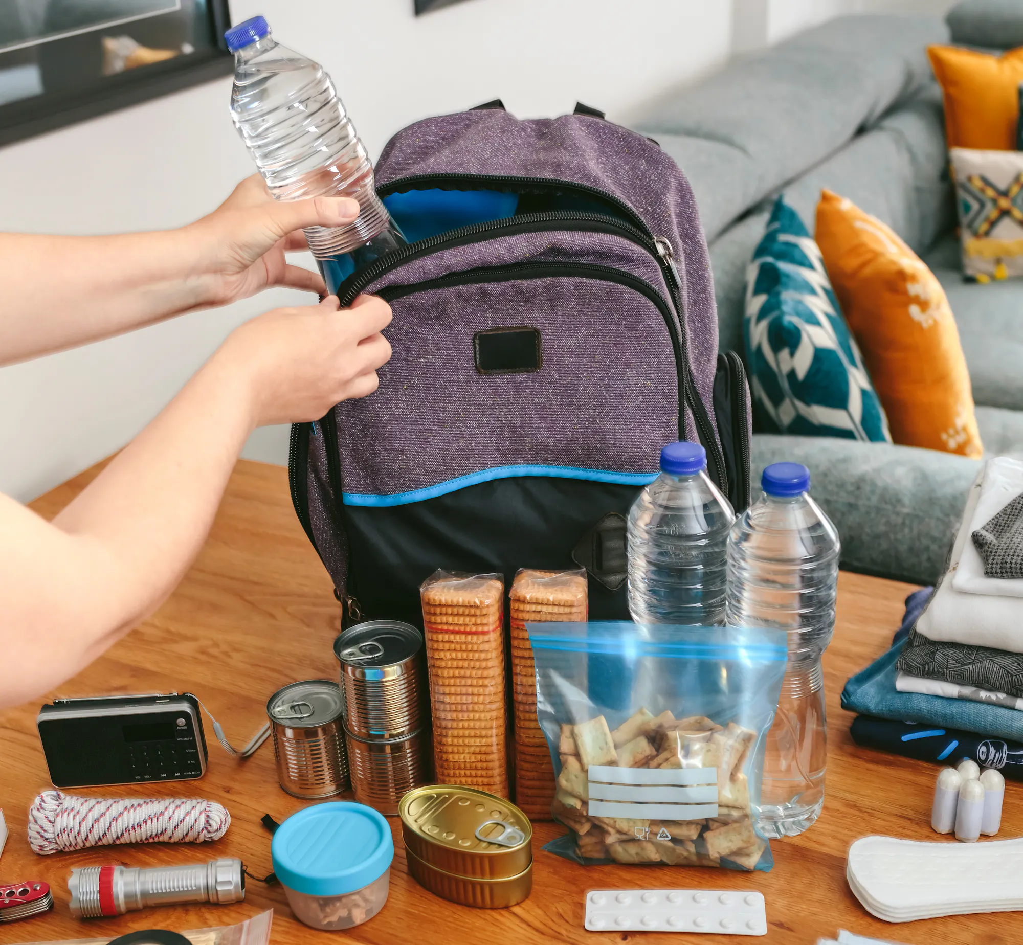 A person is packing a backpack with water bottles, food, a radio, medical supplies, and other essentials on a table.