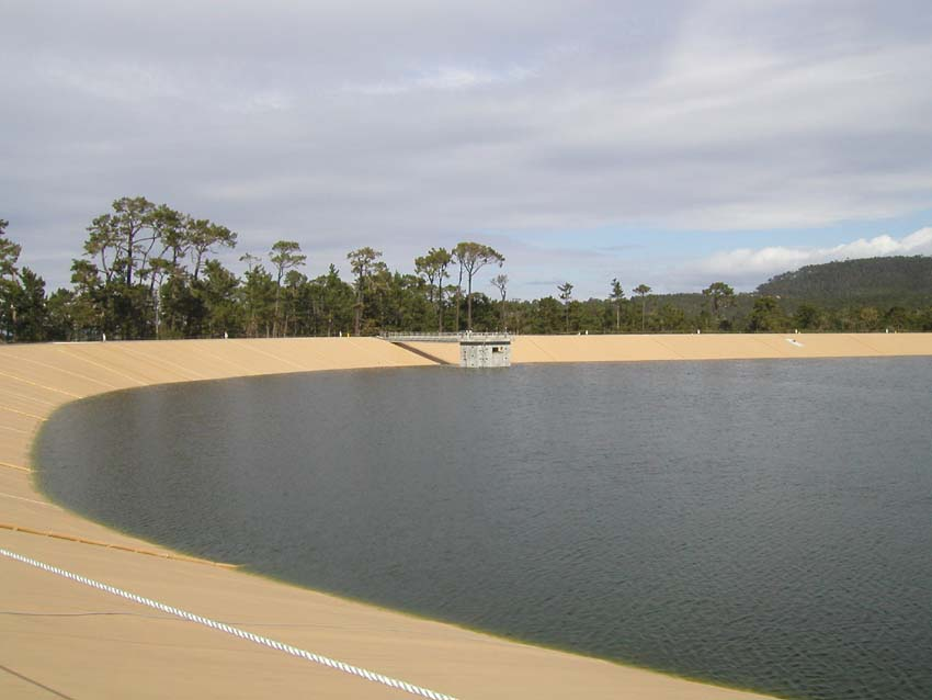 A reservoir with a sandy dam, water, trees, and a small structure, under a partly cloudy sky.