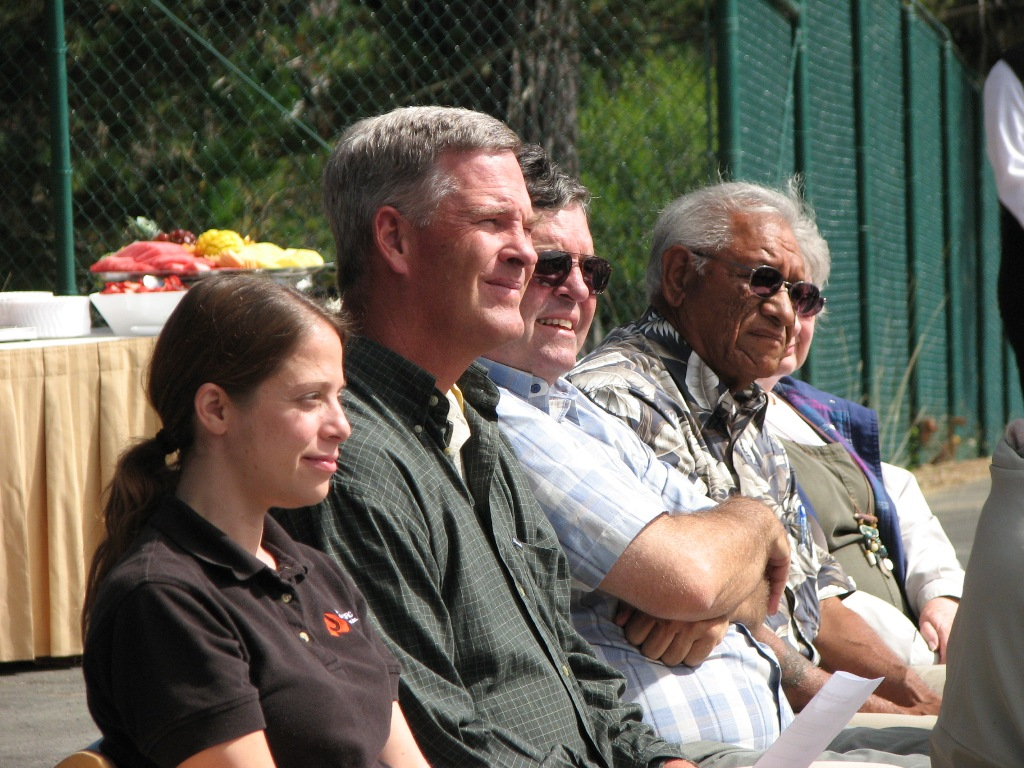 Four people sitting in a row, smiling, possibly at an outdoor event. Some wearing sunglasses; bright, sunny day.