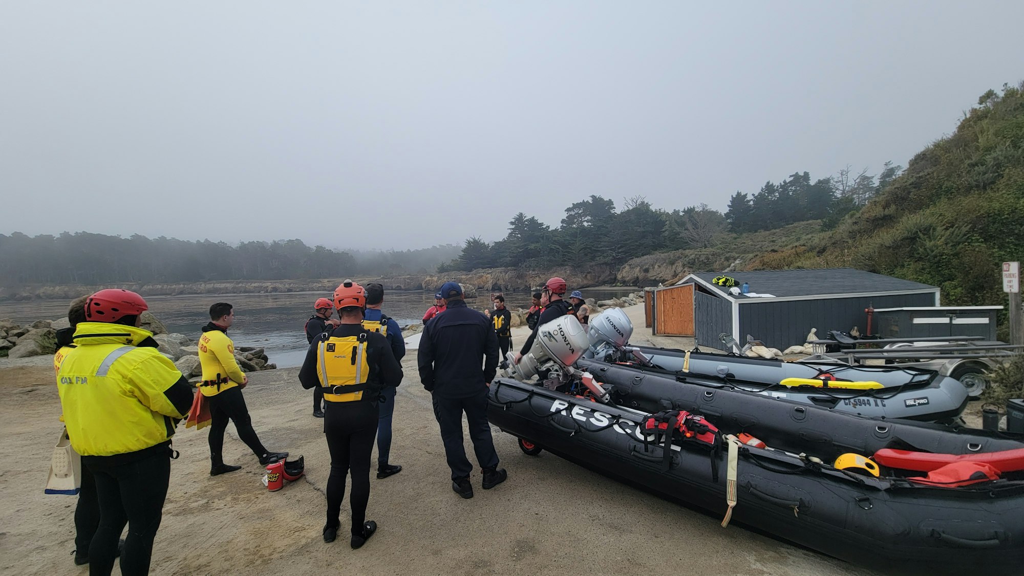 People in helmets and life vests gather by inflatable boats near a foggy shoreline.