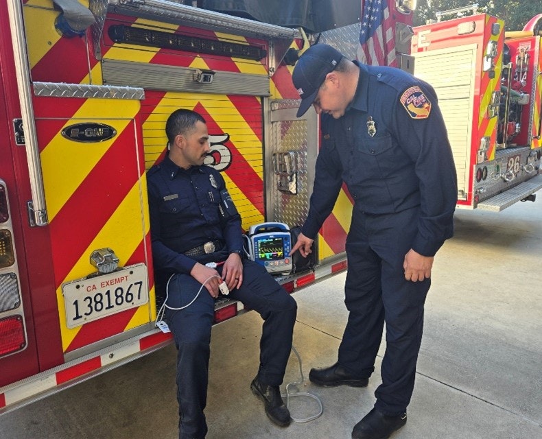 Two firefighters by a fire truck, one seated and connected to a medical monitor, the other standing and adjusting the device.
