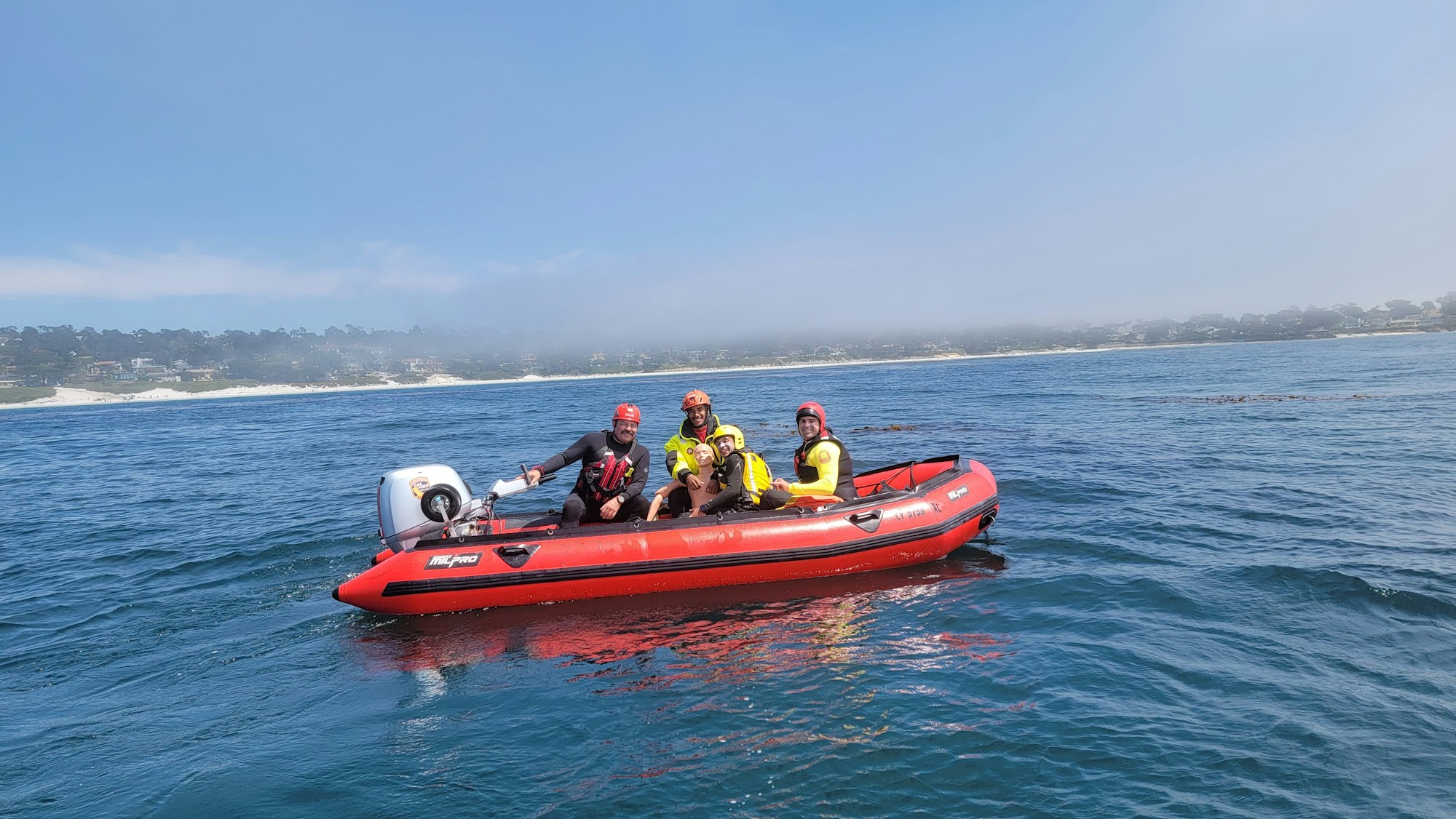 Four people in water gear on a red inflatable boat on the ocean, near a coastline.