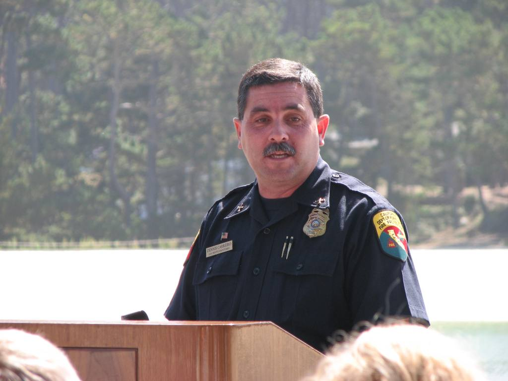 A uniformed officer standing at a podium with trees and water in the background.