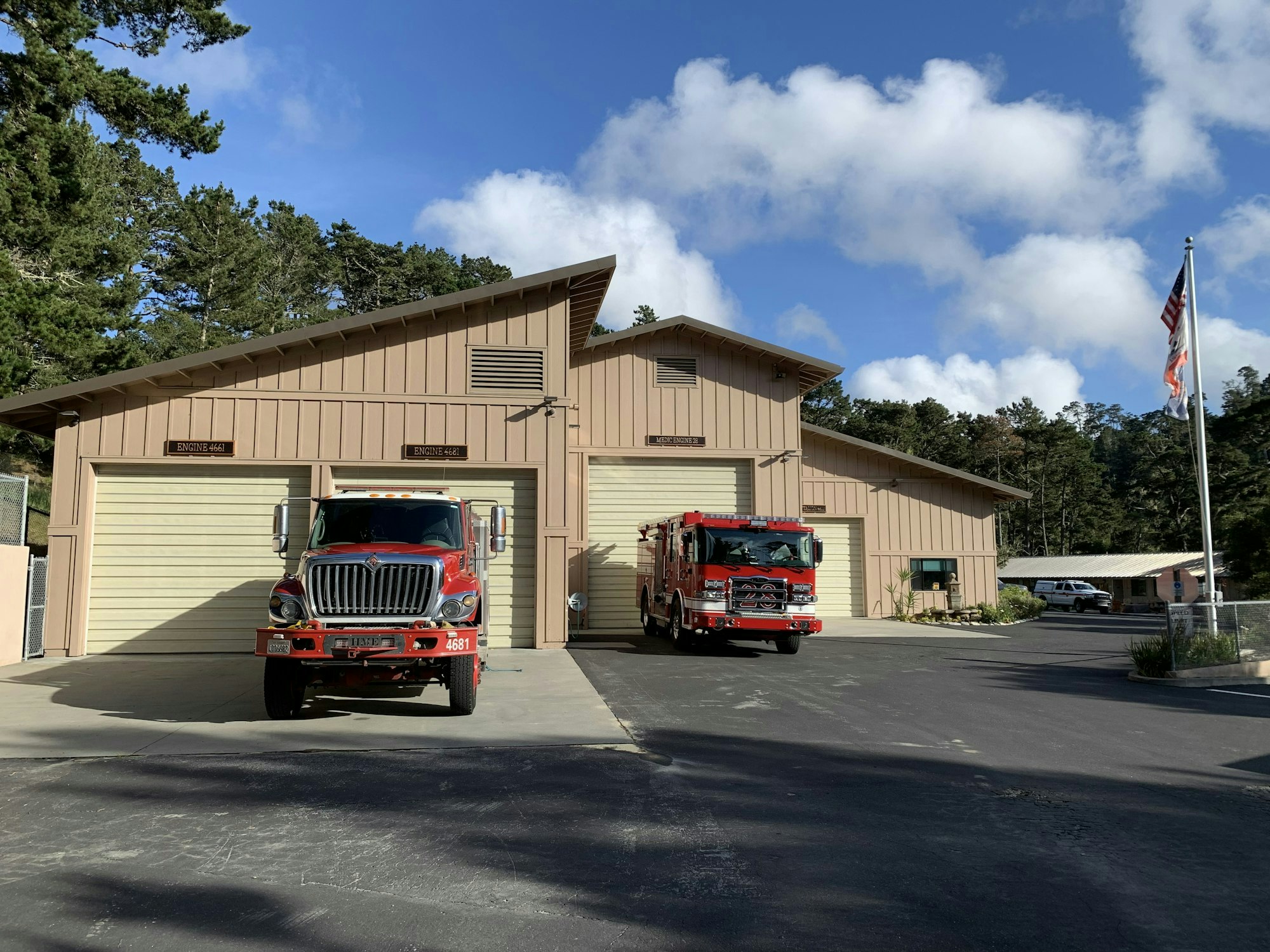 Fire station with two red fire trucks in front, surrounded by trees and a clear blue sky.