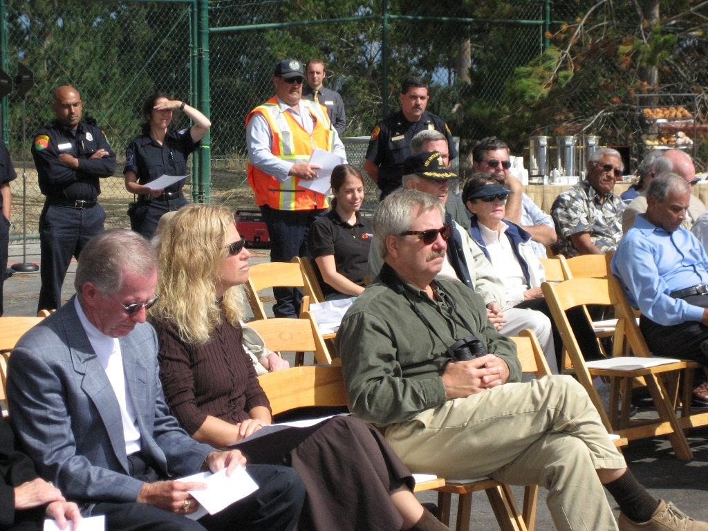 Group of people sitting at an outdoor event, with attendees in formal and work attire, and officials standing in the background.