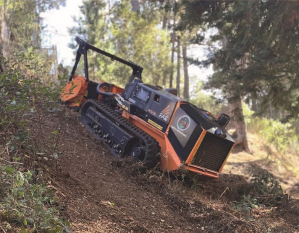 A small orange and black tracked vehicle with a clearing attachment is navigating a forested, sloped area.