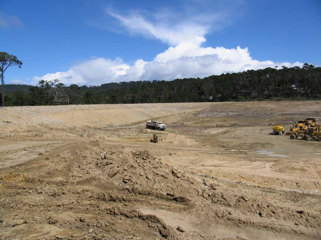 Large construction site with cleared land, heavy machinery, and a forested backdrop under a blue sky.