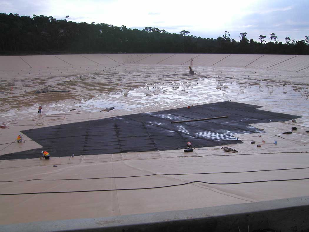 Workers on a large construction site with a vast, partially covered area by geotextile fabric, surrounded by trees.