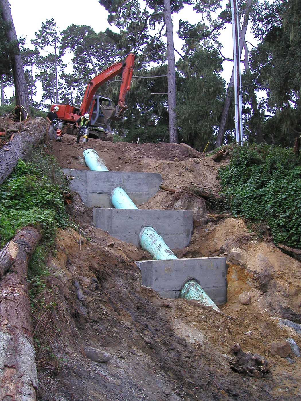An excavator on a hillside with blue pipelines and concrete structures, among trees.