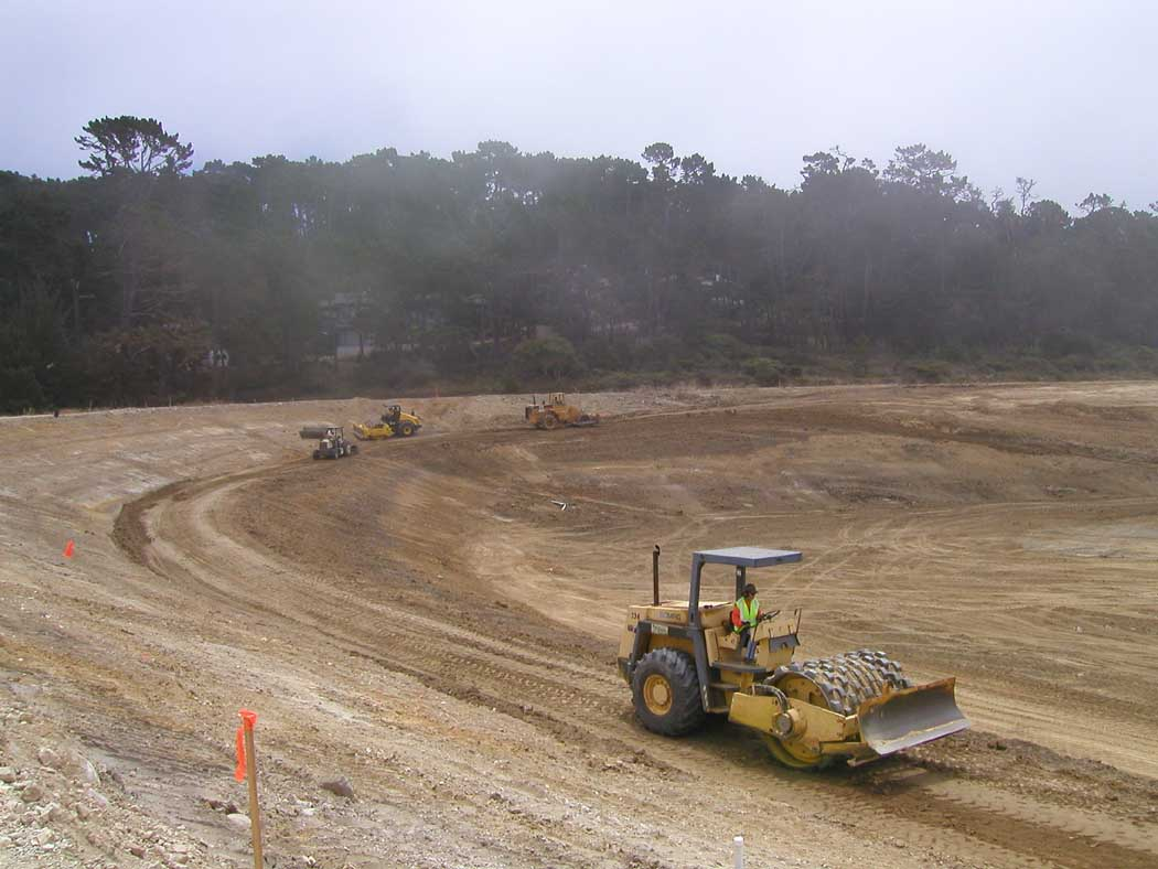 Construction site with heavy machinery and dirt roads, surrounded by trees under a hazy sky.