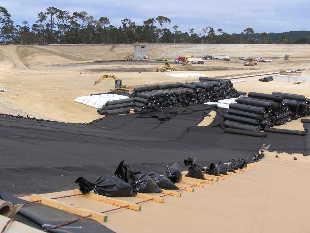 A construction site with stacked geotextile tubes, heavy machinery, and vehicles in the background.