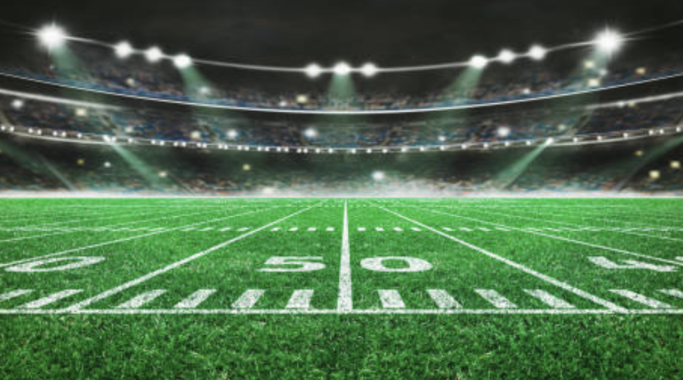 An empty football field with yard lines at night, illuminated by stadium lights.