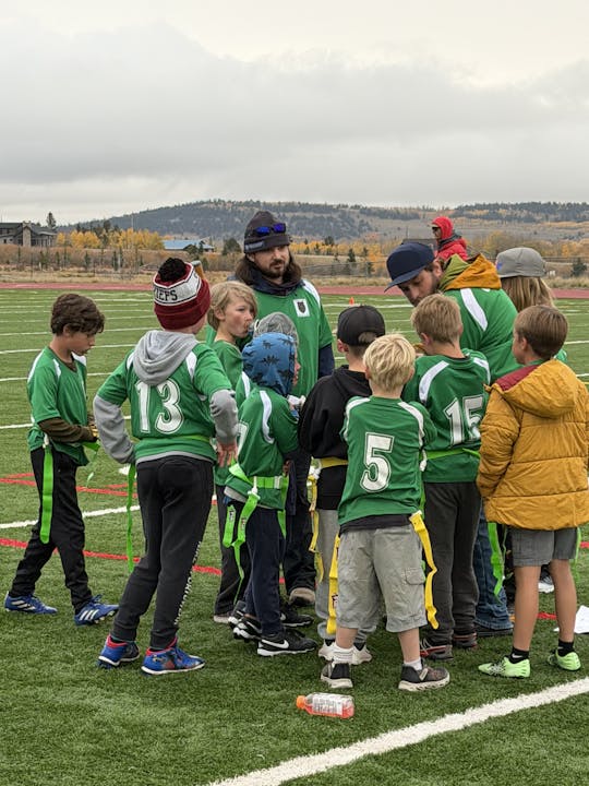 A group of kids and adults in green jerseys huddle on a sports field, listening to instructions.