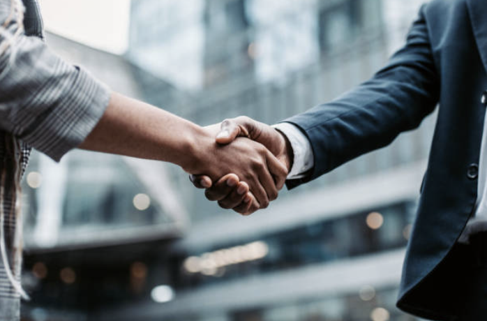 A close-up of two people shaking hands, one in a plaid shirt, the other in a suit, with a blurred urban background.