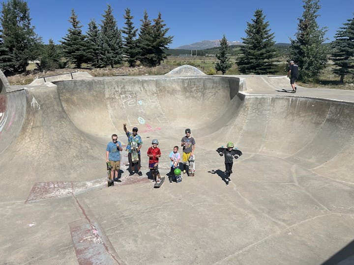 Group of skaters in a concrete skatepark bowl with trees and a mountain backdrop.
