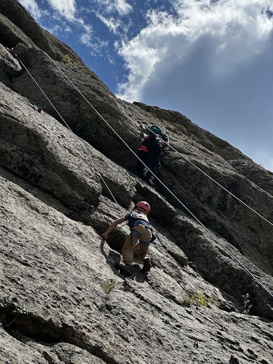 Two people rock climbing on a steep cliff, secured with ropes, under a partly cloudy sky.