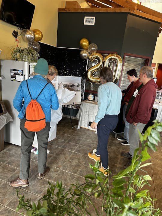 A group of people stands by a display with large "20" balloons and metallic decorations, in a tiled room.