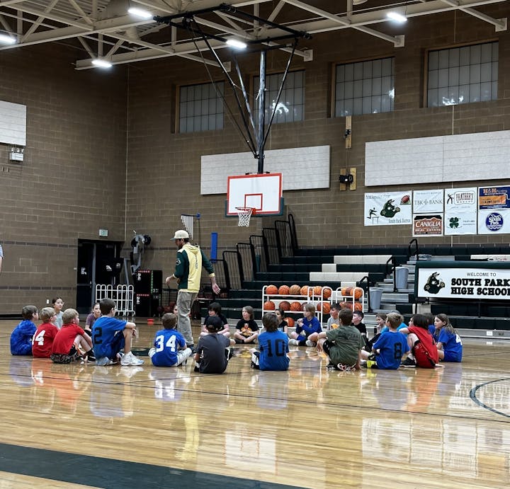 A coach addressing a group of young basketball players sitting on a gym floor.