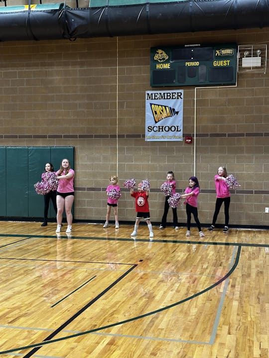 A group of children in a gymnasium holding pink pom-poms, standing in front of a scoreboard and school banner.